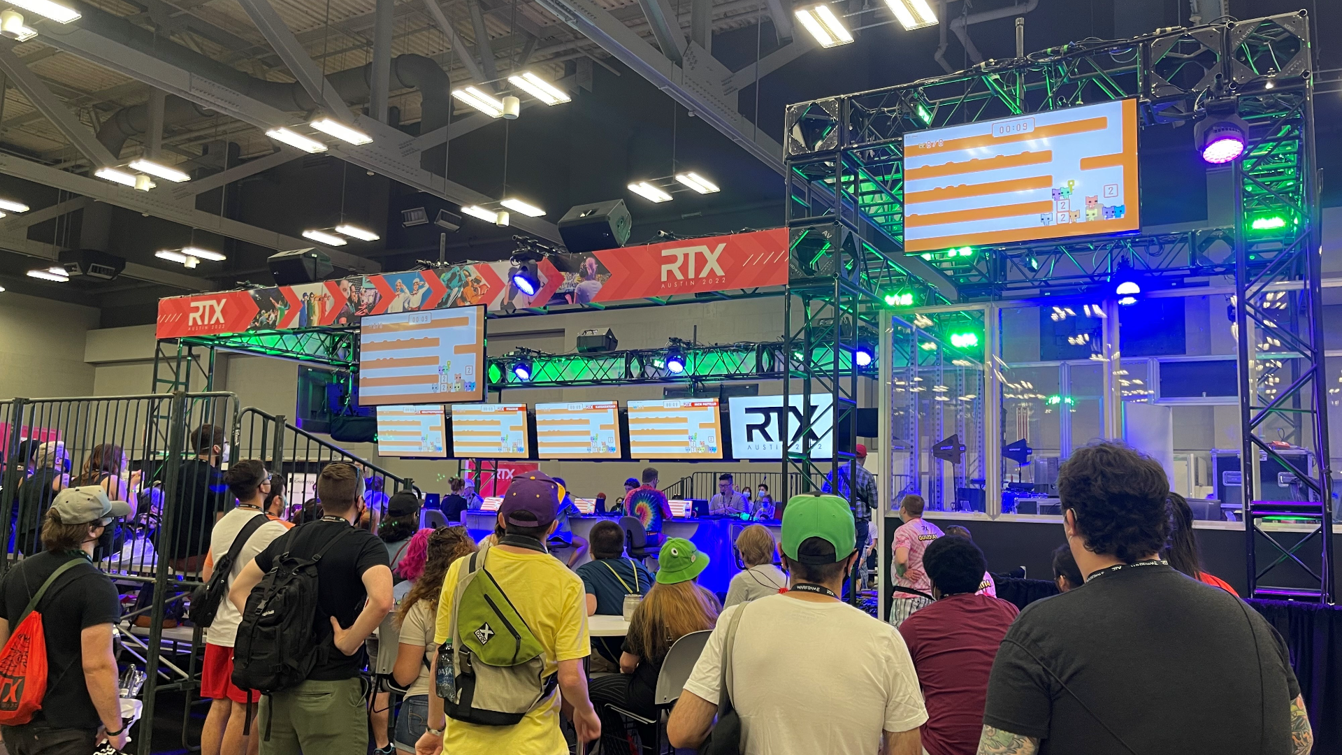 A shot of RTX Austin's Gaming Pavilion. Above the crowd of people are several television screens displaying visuals of the game "PICO PARK", and above that is a banner for RTX showing several images from various RT Productions.